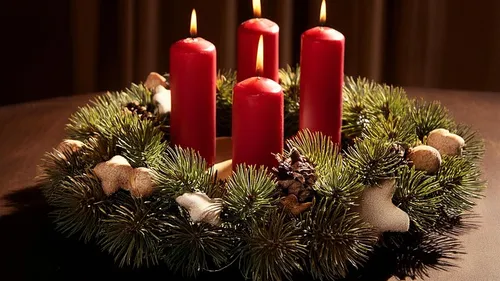 Four red candles on green wreath, lit, indoors.