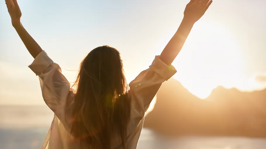 Person raising arms towards sunrise on beach.