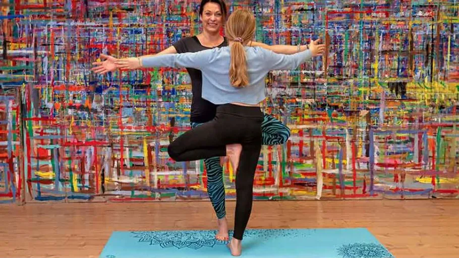 Zwei Frauen machen Yoga drinnen, bunter Hintergrund.