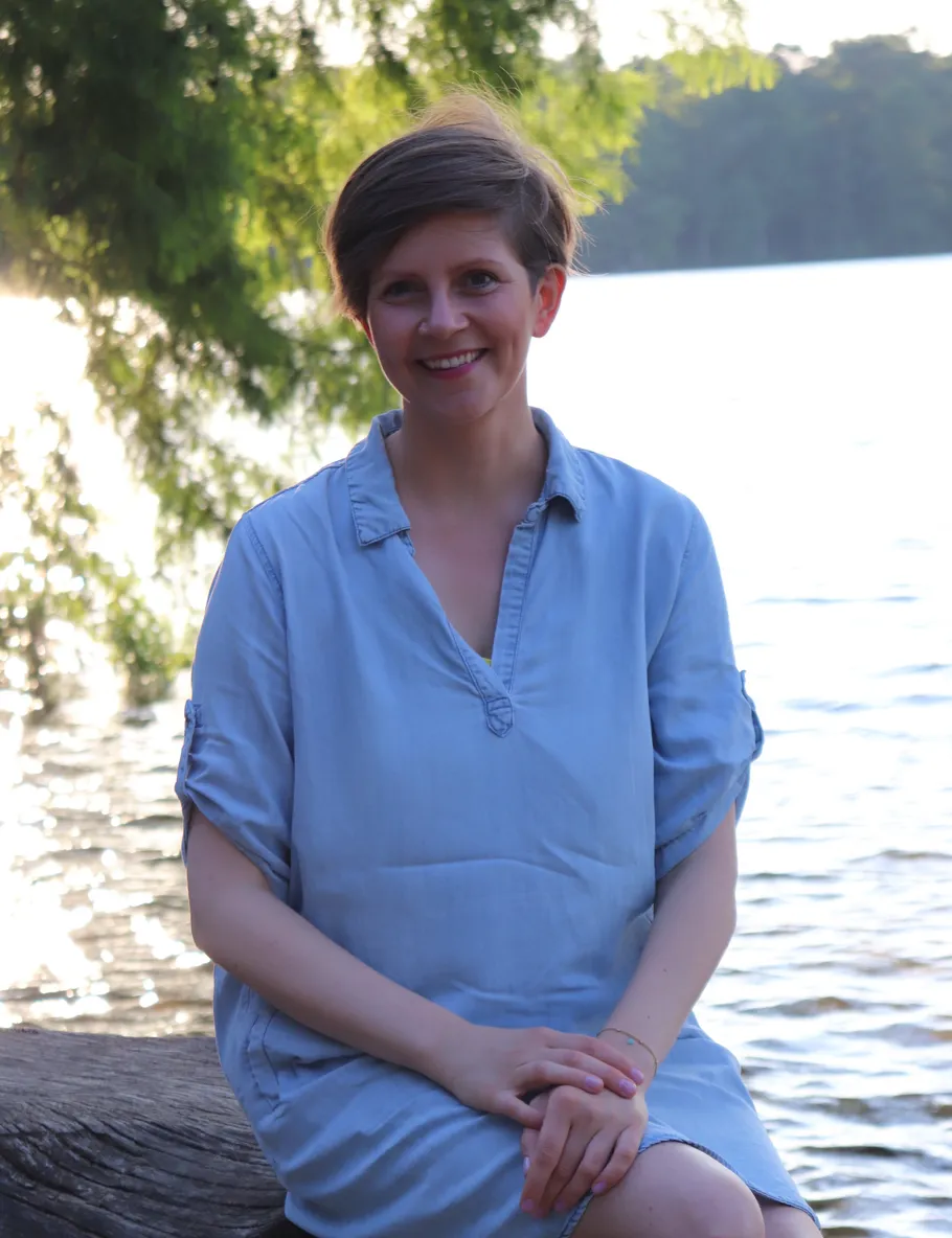 Smiling person sitting by lakeside, trees in background.