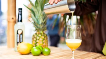 Person gießt Cocktail nahe Früchten und Flasche.