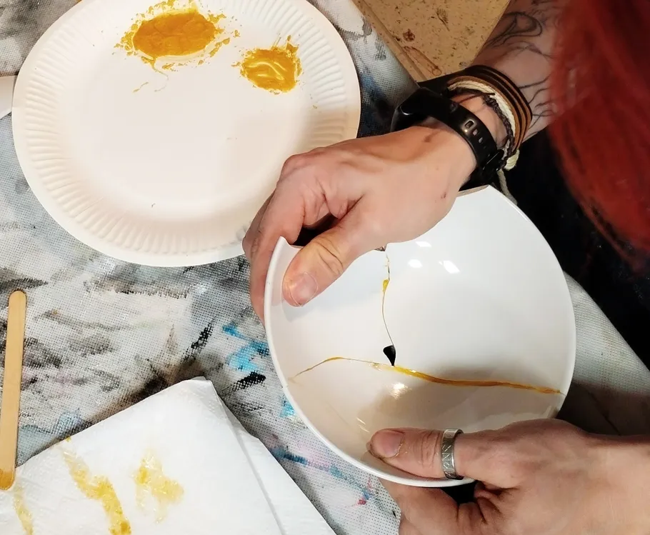 Hände reparieren eine zerbrochene Schüssel mit Goldfarbe.
