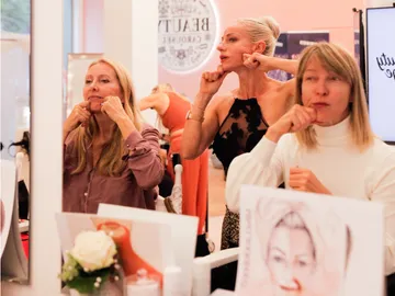Three women practice facial massages in front of mirrors in a beauty studio.