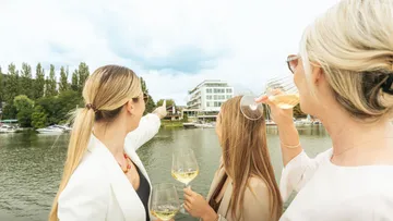 Women with wine glasses by riverside building.