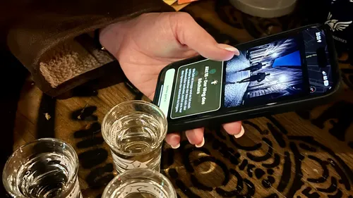 Hand hält Handy mit Bildern, neben Gläsern.