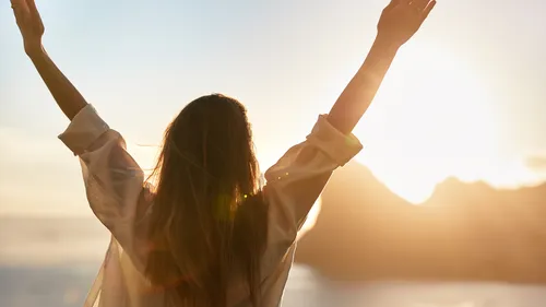 Woman raising arms towards sunrise outdoors.