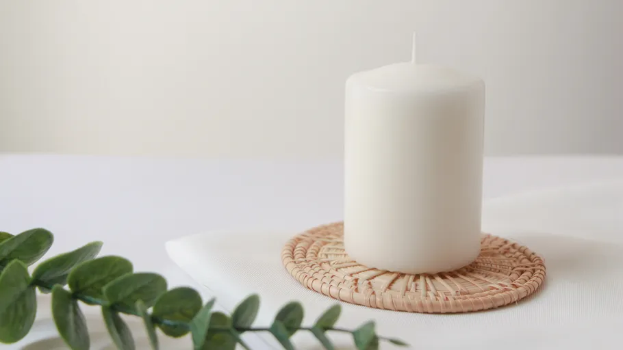 White candle on woven coaster, leafy background.