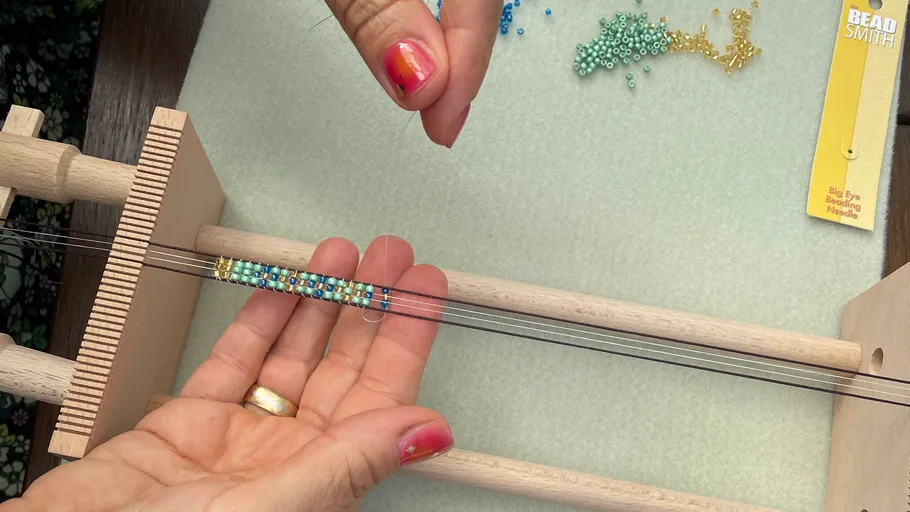Hands weaving beads on a wooden loom.