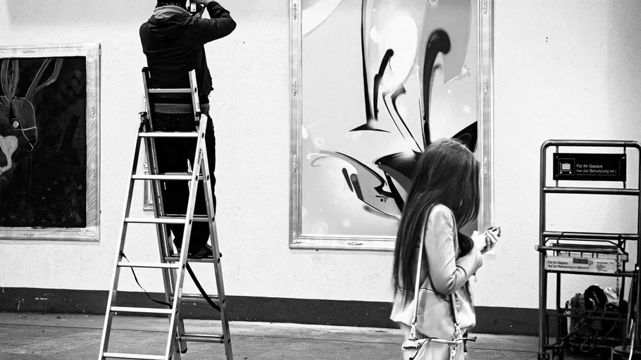 Mann auf Leiter fotografiert Kunstwerk; Frau läuft vorbei.