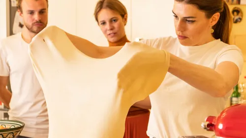 Woman stretching dough in kitchen with onlookers.