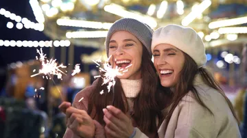 Two women laughing, holding sparklers, nighttime fair.