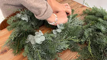 Hände arrangieren einen Kranz auf Holztisch.