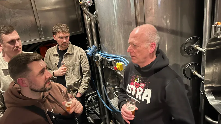 Men talking near brewery equipment, holding drinks.