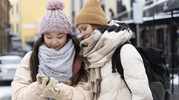 Two women bundled up looking at a phone outside.