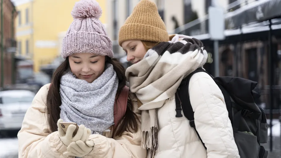 Two women in winter clothing look at a phone outdoors.