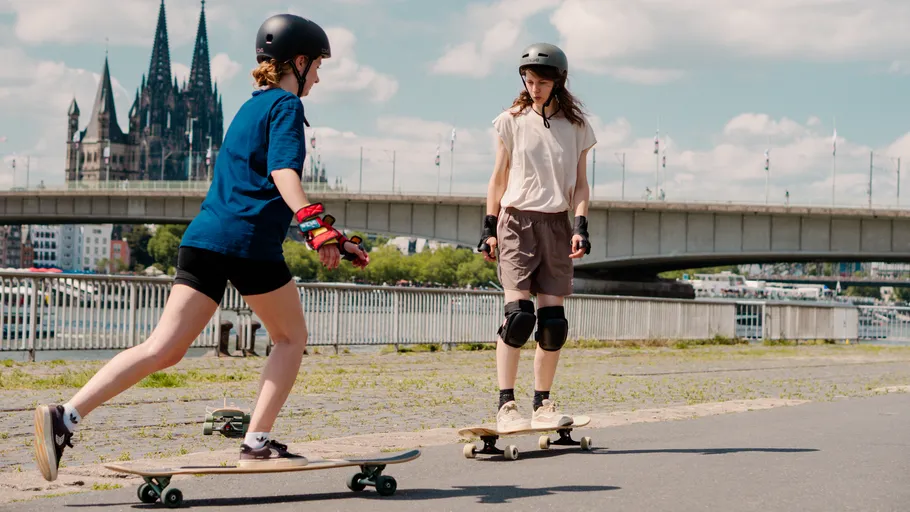 Two skateboarders cruising near a city bridge.