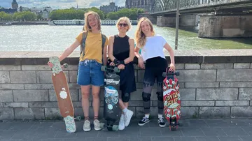 Three women with longboards by a riverside.
