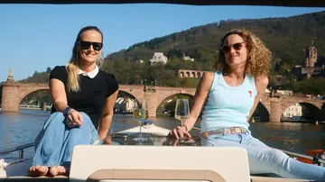 Two people sitting on a boat, bridge behind.