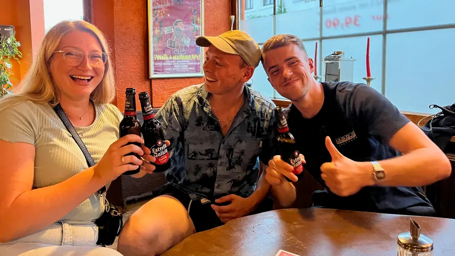 Three people laughing, clinking beer bottles indoors.