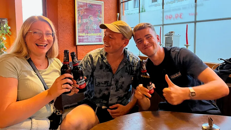 Three people smiling, holding beer bottles, indoors.