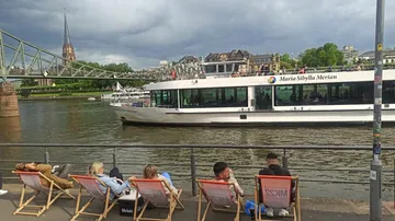 People relax on chairs, boat passes on river.