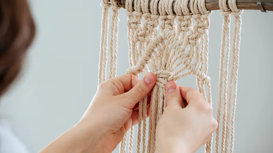 Hands knotting macramé ropes on wooden rod.