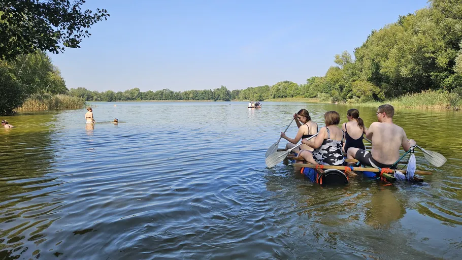 People on raft paddling across wooded lake.