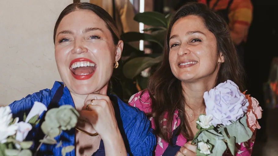 Two women smiling, holding flowers indoors.