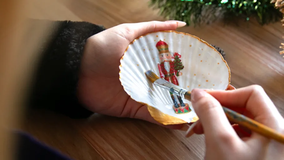 Person painting nutcracker on seashell indoors.