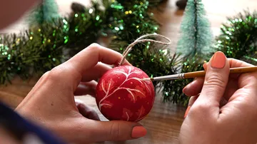Person painting red ornament with brush, greenery nearby.