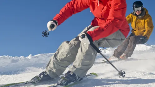 Zwei Skifahrer fahren verschneite Abfahrt unter blauem Himmel.