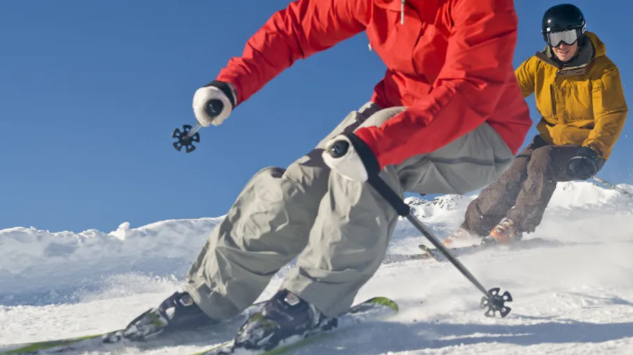 Zwei Skifahrer fahren verschneite Abfahrt unter blauem Himmel.