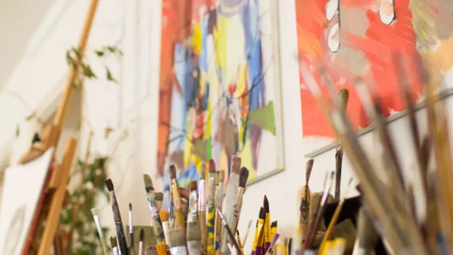 Paintbrushes in focus, colorful paintings behind.
