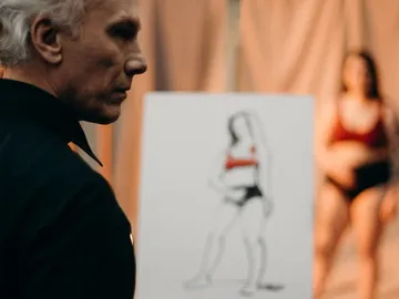 Man looks at a sketch of a posing woman in the background.