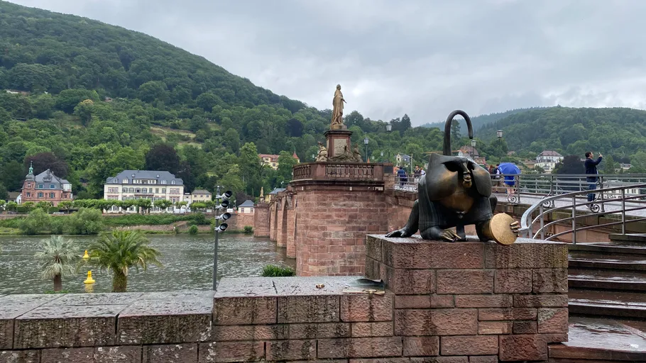 Skulptur nahe einer historischen Brücke und grünen Hügeln.