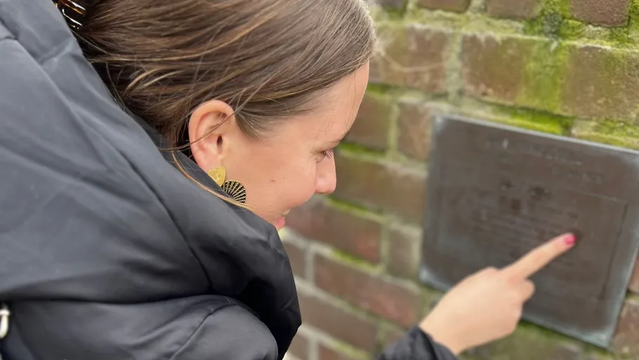 Person zeigt auf eine Plakette an Ziegelwand.