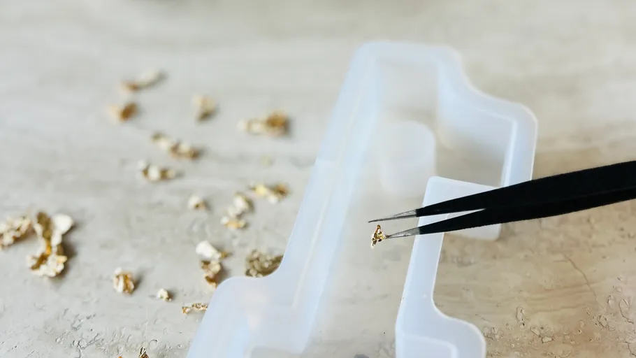 Tweezers holding small gold nugget over container.