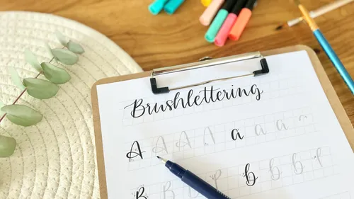 Brushlettering-Übungsblatt auf Klemmbrett, Stifte daneben.