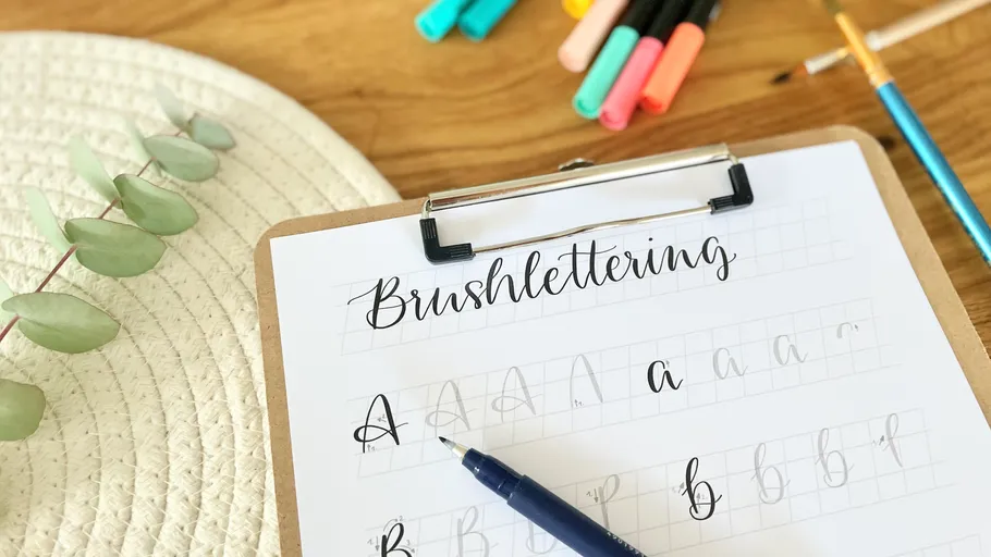 Brush lettering practice sheet, pens on wooden table.