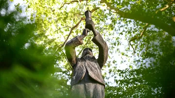Statue holding torch amidst green trees.
