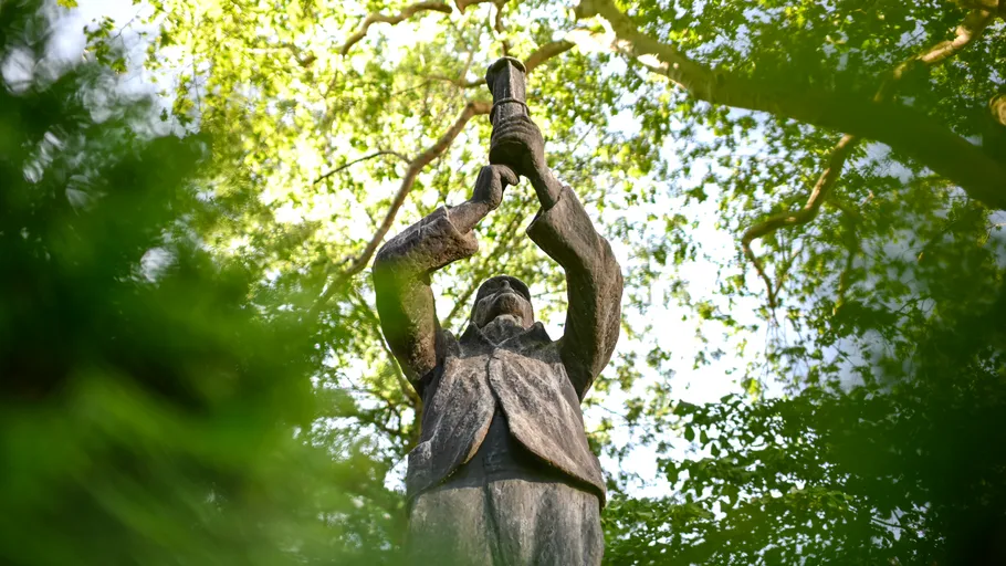 Statue hält Fackel zwischen grünen Bäumen.