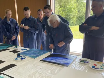 Group of people watch elderly woman painting.