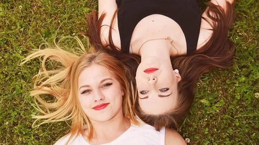 Two women lying on grass, heads touching.