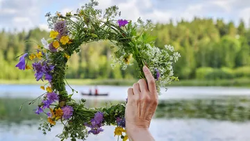 Hand holds flower wreath by a lake.