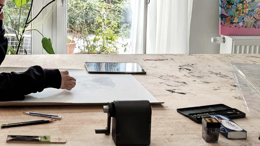 Person zeichnet auf Papier, Tisch mit Kunstbedarf.