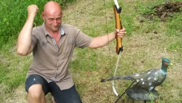 Mann kniet mit Bogen neben Zielvogel.