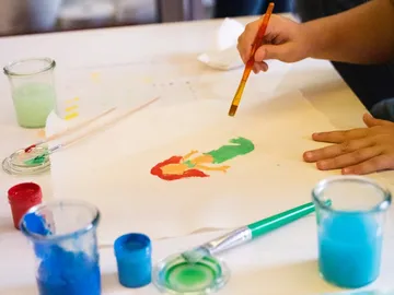 A child paints with a brush on paper, surrounded by colors.