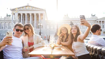 Group taking selfies at outdoor cafe, parliament visible.