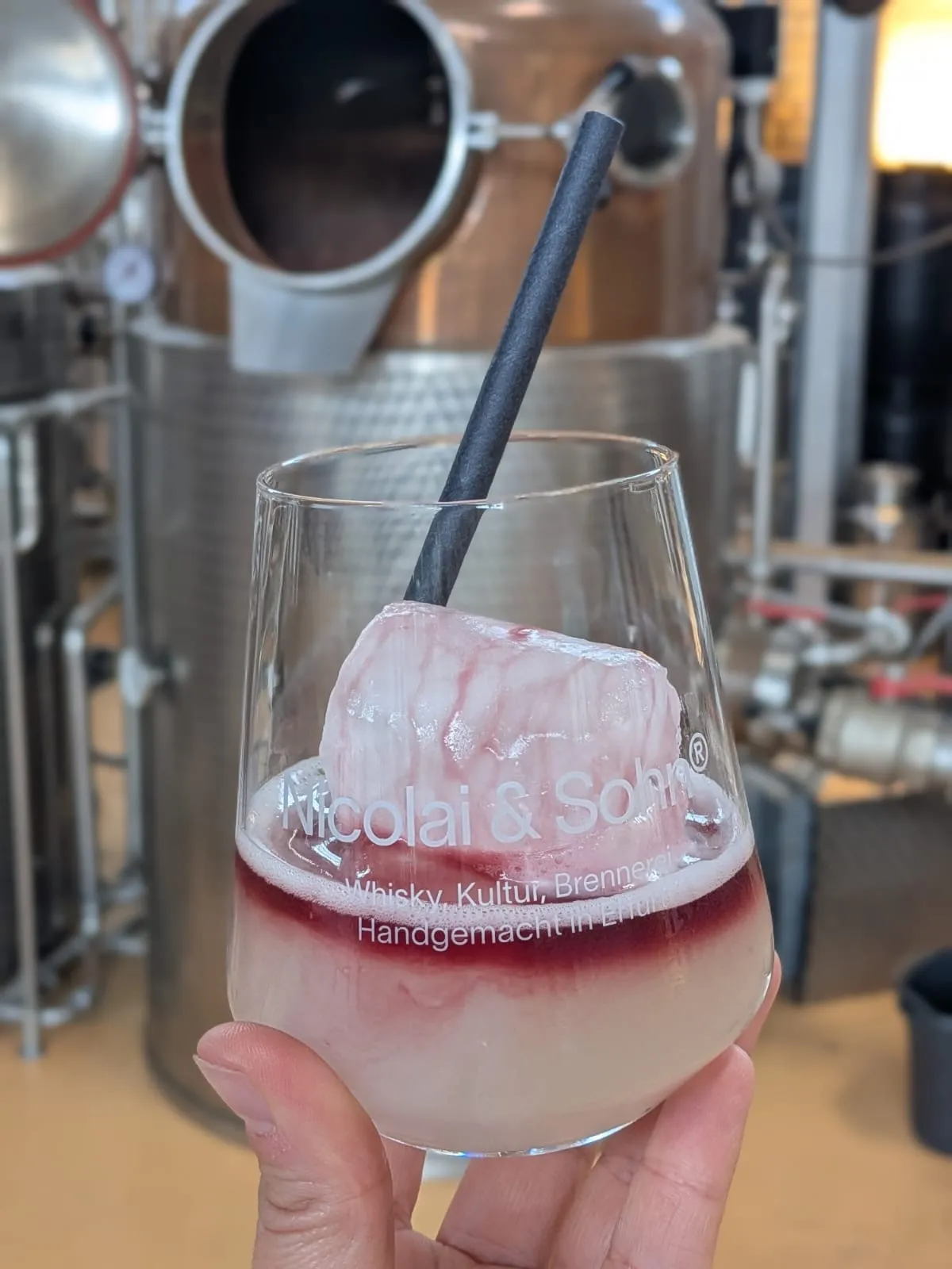 Hand hält Cocktail vor Destilleriehintergrund.