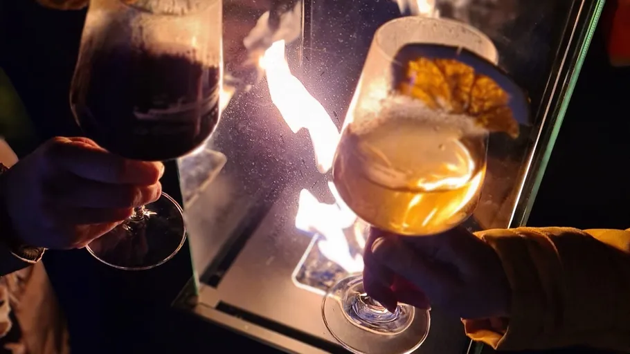 Zwei Hände stoßen Weingläser am Feuer an.